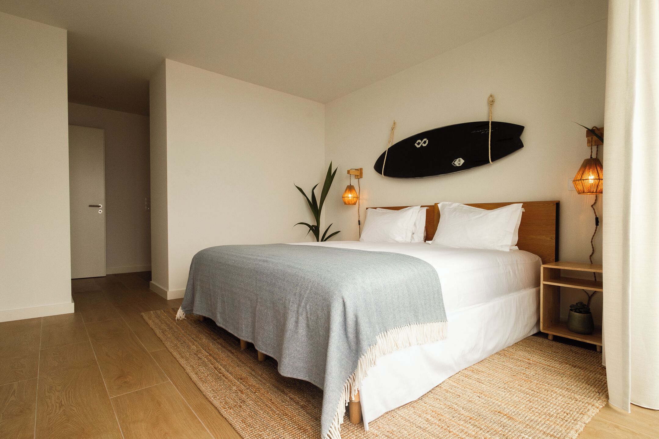 A bedroom with a bed, a surfboard on the wall, a plant, and a nightstand with a lamp.