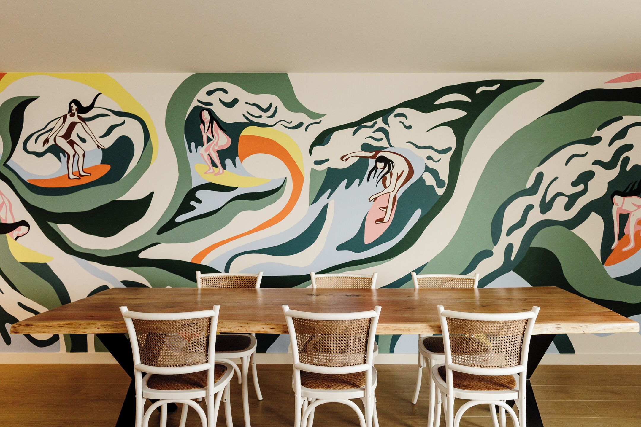 A dining area with a wooden table and wicker chairs, set against a colorful abstract mural on the wall.