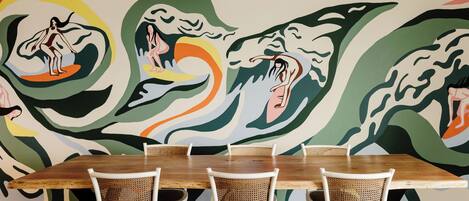 A dining area with a wooden table and wicker chairs, set against a colorful abstract mural on the wall.