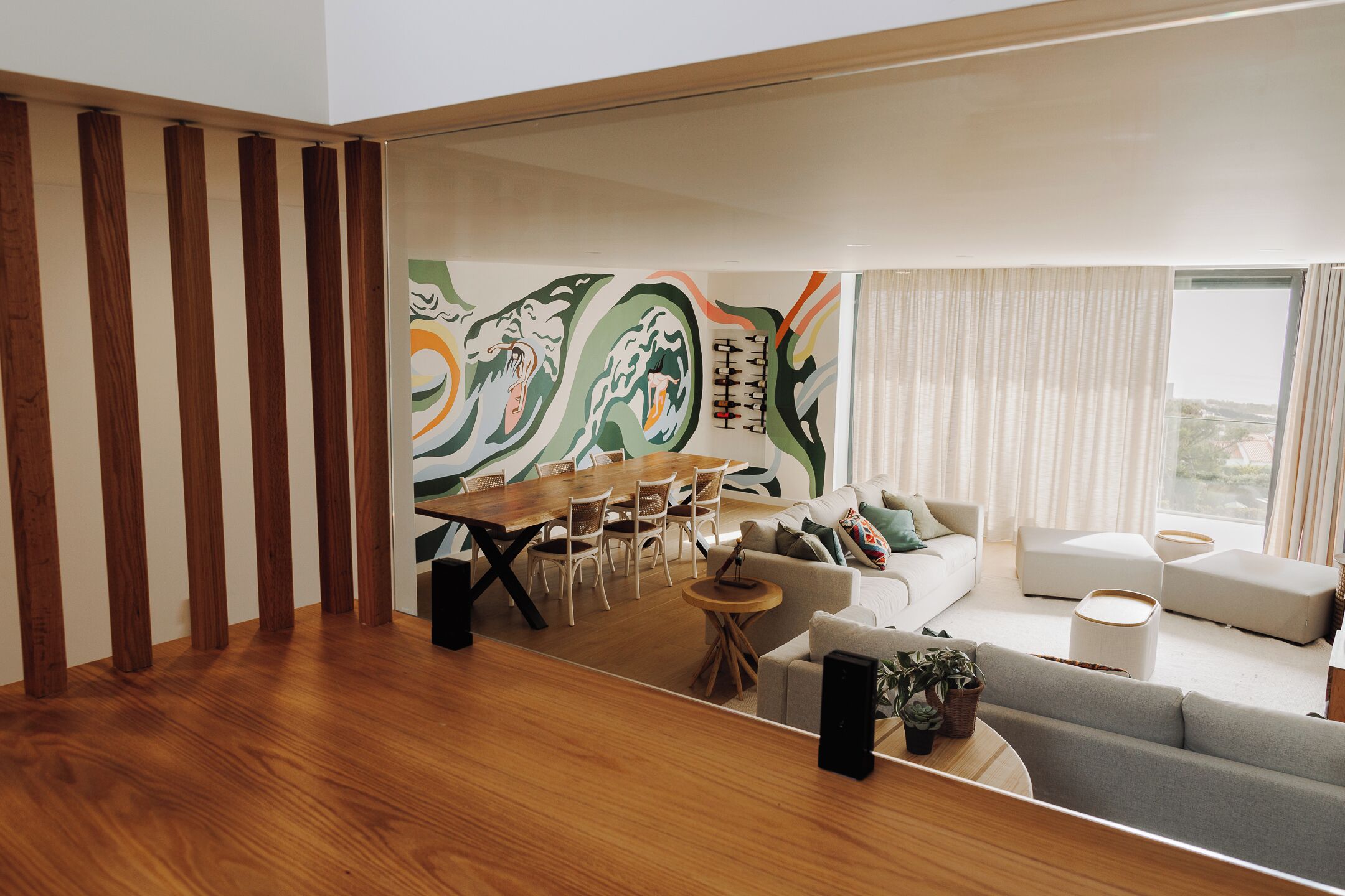 Una sala de estar moderna con piso de madera, un comedor con mesa y sillas de madera, un sofá gris y un ventanal con cortinas.
