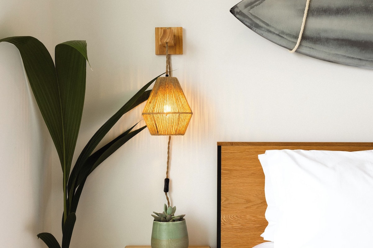 A neatly arranged bedroom with a bed, a bedside lamp, a small wooden nightstand, a potted plant, and a wall-mounted surfboard.