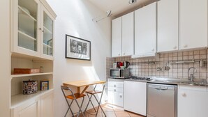 Una cocina compacta con armarios blancos, una pequeña mesa de comedor, una silla plegable, un microondas, una tetera y un lavavajillas.