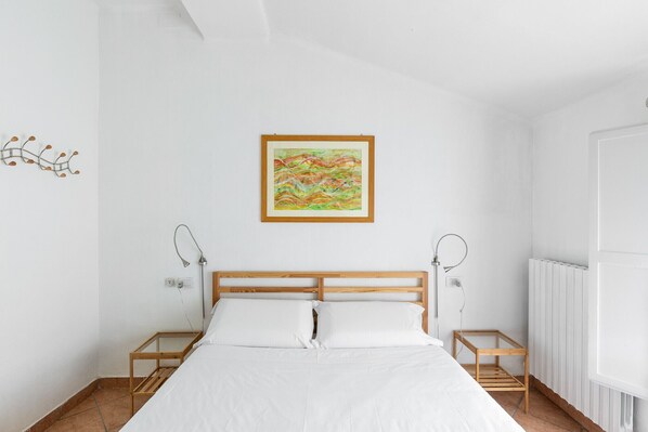 Un dormitorio con cama de madera, ropa de cama blanca, un cuadro abstracto colorido, lámparas de noche y un radiador.