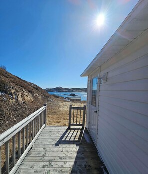 A wooden deck leading to a white building with a view of the ocean and hills. - Salty B'y Cottage (Twillingate)