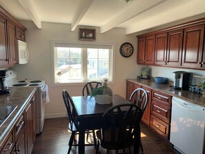 A kitchen with wooden cabinets, a dining table with chairs, a clock, and a window with a view of the outside.