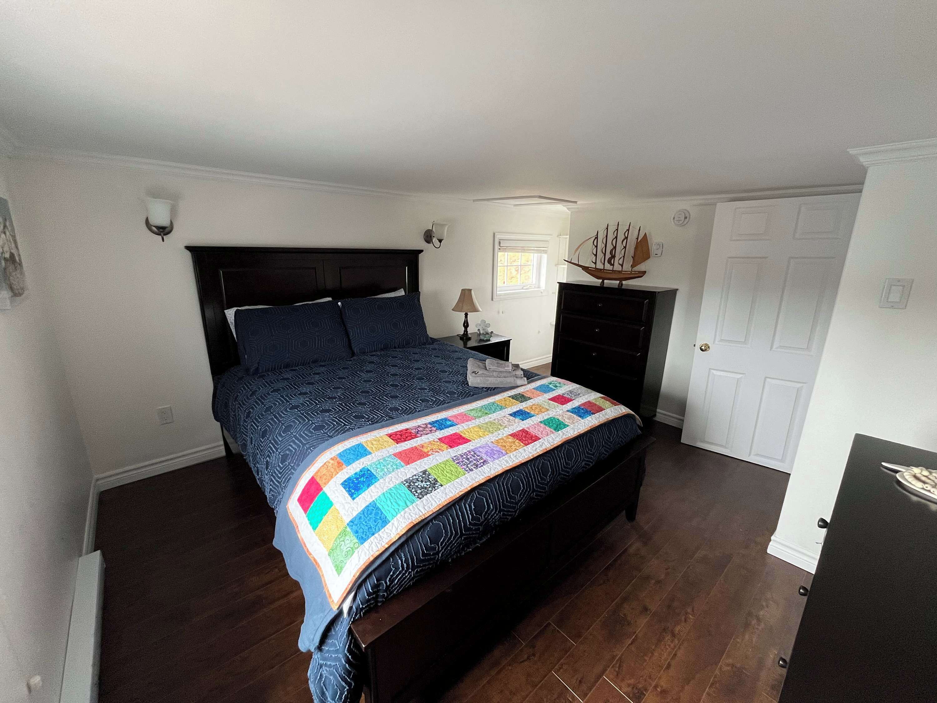 Een slaapkamer met een donker houten bed, een kleurrijke quilt, een nachtkastje met een lamp, een dressoir en een deur.