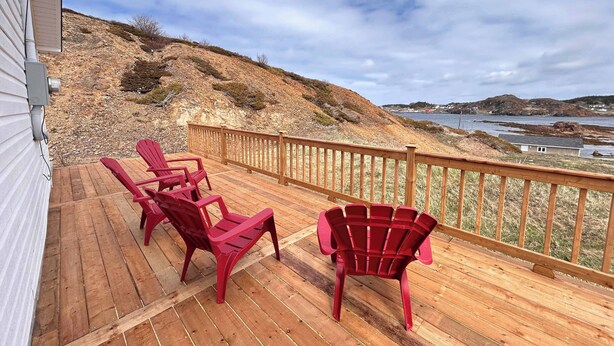 A wooden deck with red chairs and a railing overlooking a coastal landscape. - Salty B'y Cottage (Twillingate)