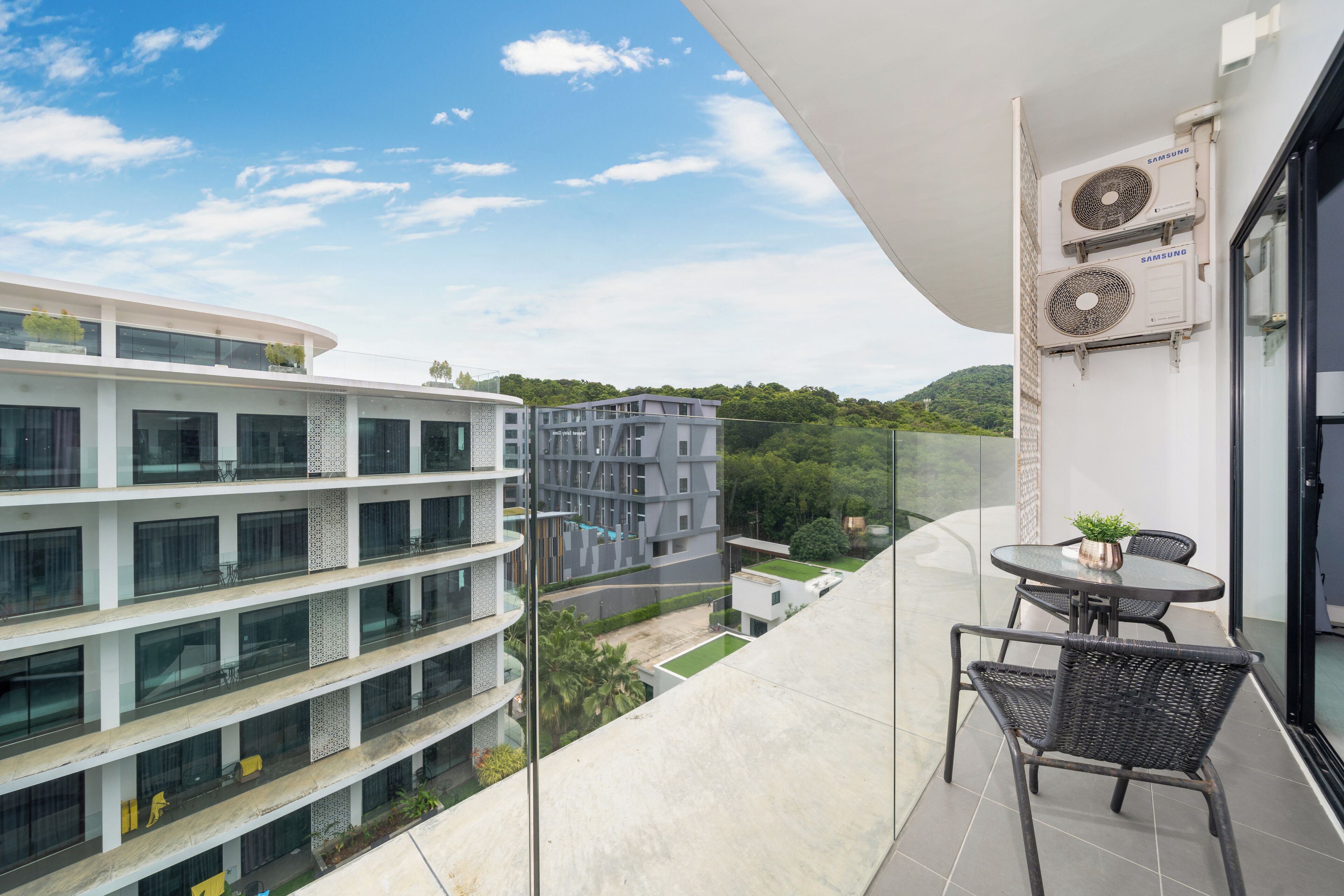 A balcony with a table and chairs, overlooking a cityscape with modern buildings and a green hill in the distance.