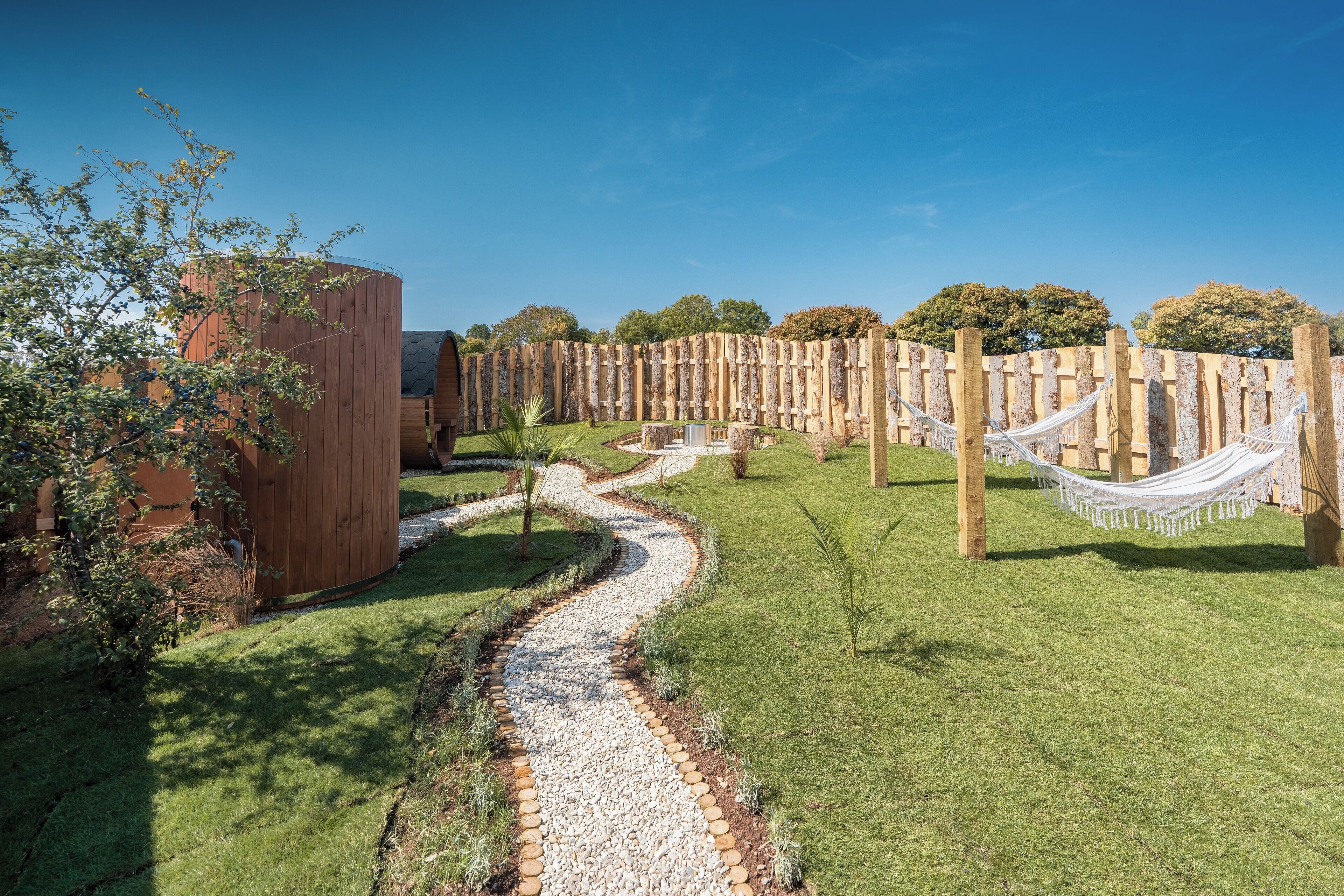 A garden pathway with a wooden fence, a hammock, and a building with a sloped roof.