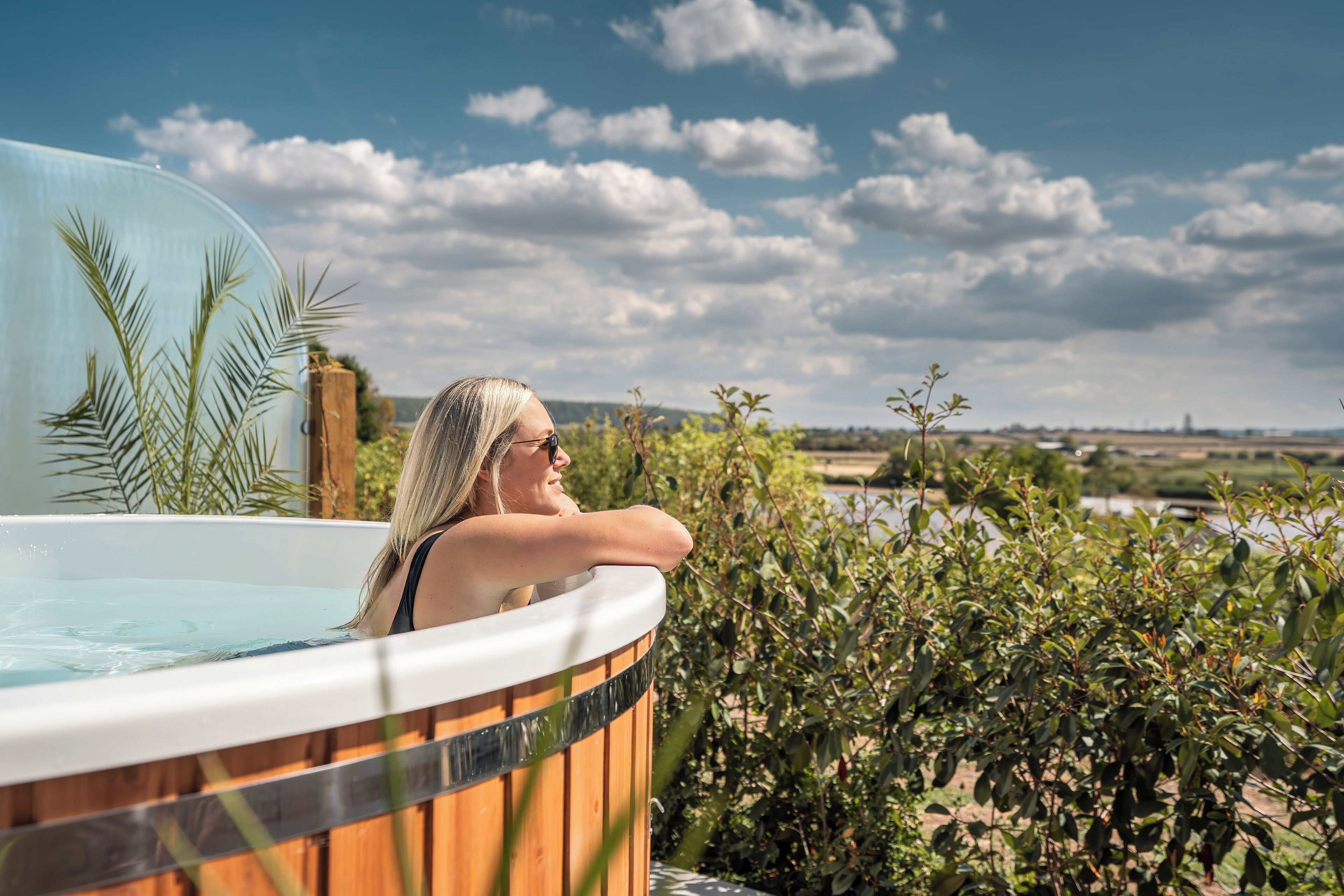 A person relaxing in a wooden hot tub with a view of a body of water and greenery.