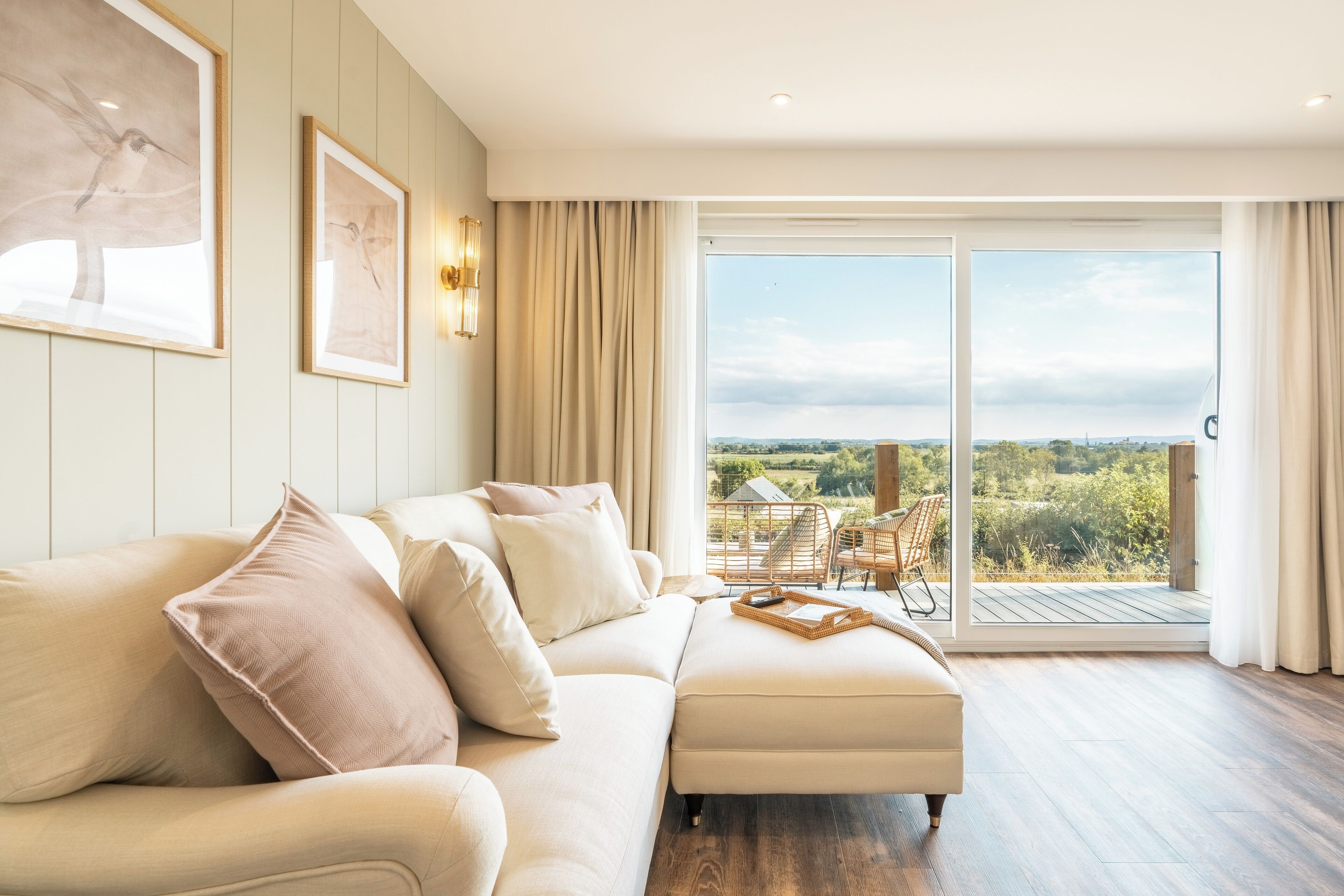 Ein modernes Wohnzimmer mit einem beigefarbenen Ecksofa, einem Holztisch und einer großen Schiebeglastür, die auf einen Balkon mit Blick auf Grünflächen und eine entfernte Landschaft führt.