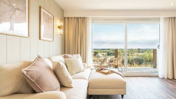 Ein modernes Wohnzimmer mit einem beigefarbenen Ecksofa, einem Holztisch und einer großen Schiebeglastür, die auf einen Balkon mit Blick auf Grünflächen und eine entfernte Landschaft führt.