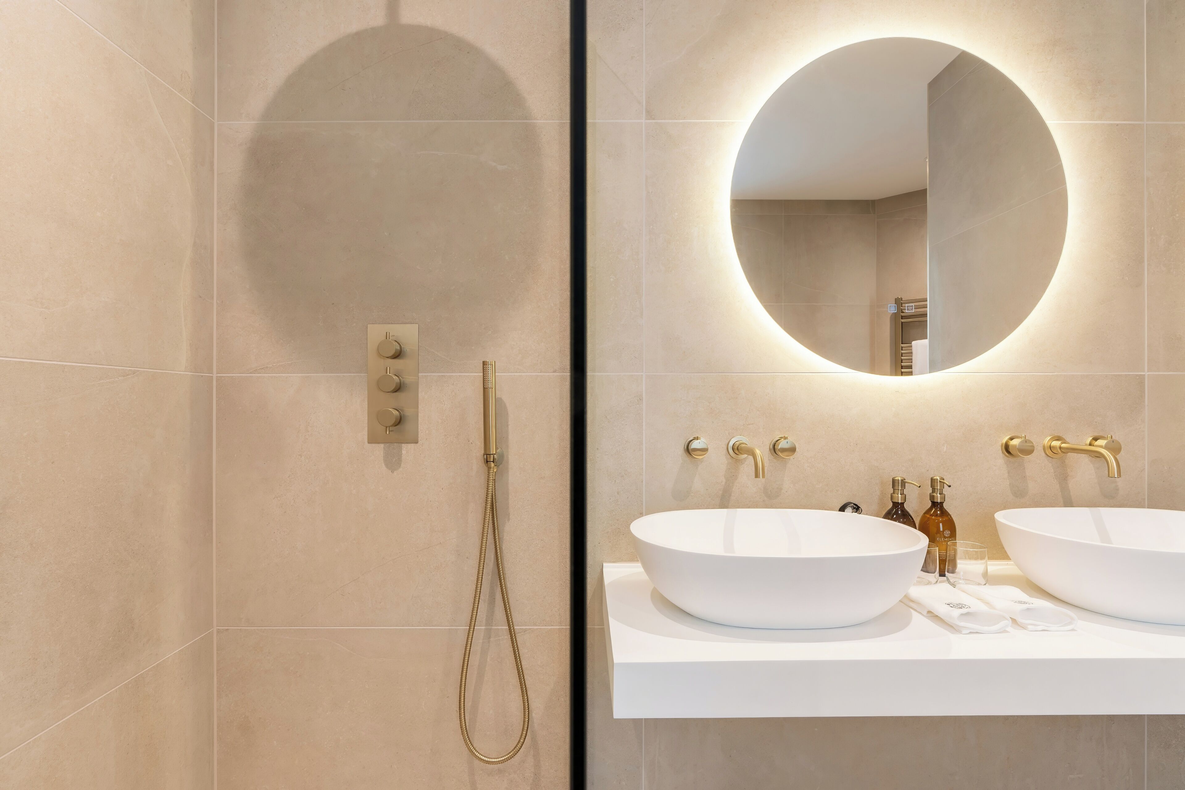 A modern bathroom with two large white sinks, a round mirror with integrated lighting, and a handheld showerhead.