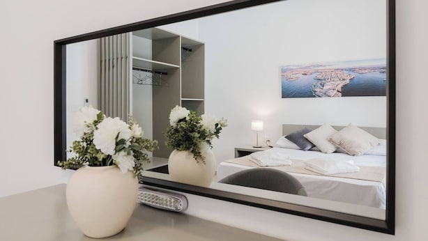 Una habitación de hotel con un espejo grande, una cama con ropa de cama blanca, una mesita de noche con una lámpara y un cuadro de un paisaje urbano costero.