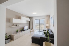 A modern living room with a sofa, a coffee table, and a TV. There are plants and a balcony with a view.