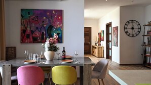 Un comedor con una mesa dispuesta para cuatro personas, un cuadro abstracto grande encima de la mesa y un reloj en la pared.