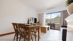 Un comedor con mesa y sillas de madera, una mesita auxiliar, un televisor y un balcón con vistas al paisaje.