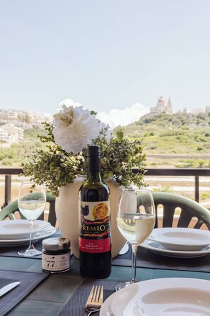 A table set for dining with a bottle of red wine, a vase with flowers, and a scenic view of a hill with buildings in the background.