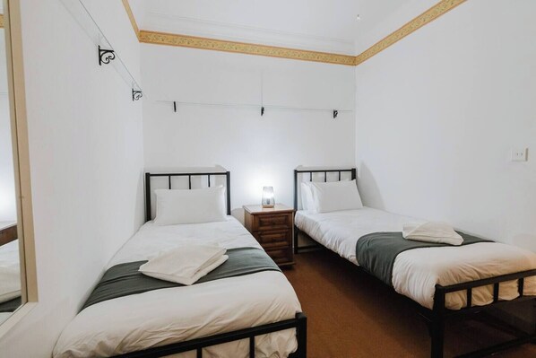 Habitación con dos camas individuales, cada una con estructura de madera y ropa de cama blanca. Entre las camas, hay una pequeña mesita de noche con una lámpara. Las paredes son blancas y el techo tiene molduras de madera.