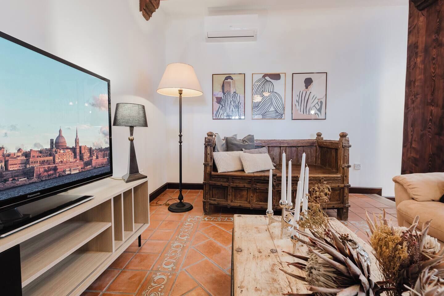 Una sala de estar con televisor, una lámpara de pie, un banco de madera con cojines, una mesa de centro con objetos decorativos y tres obras de arte enmarcadas en la pared.