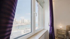 Une chambre dotée d’une grande fenêtre donnant sur le bord de l’eau, d’un lit, d’une table de chevet avec une lampe et d’un rideau.