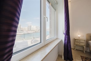 A room with a large window overlooking a waterfront, a bed, a nightstand with a lamp, and a curtain.