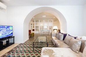 Una sala de estar con sofá, televisor, mesa de centro, alfombra estampada y un arco que da a un comedor.