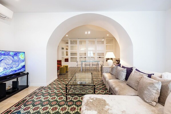 A living room with a sofa, TV, coffee table, patterned carpet, and an arched doorway leading to a dining area.