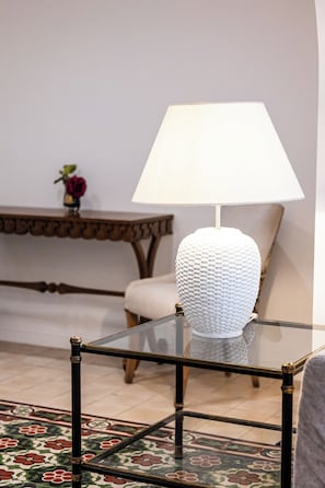 Una mesa con superficie de vidrio, una lámpara blanca con textura, una silla y una mesita con un florero de flores.