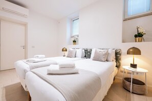 Habitación de hotel con dos camas, una mesita de noche con lámpara, un ventanal pequeño y una puerta.