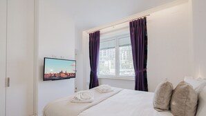 Une chambre d’hôtel avec un lit, un téléviseur fixé au mur, une fenêtre avec des rideaux et un tableau représentant un paysage urbain accroché au mur.
