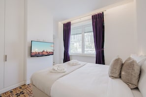 A hotel room with a bed, a television mounted on the wall, a window with curtains, and a cityscape picture on the wall.