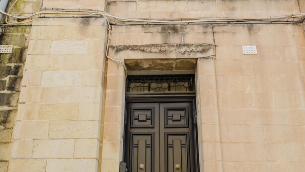 Un bâtiment doté d’une porte à deux battants de couleur brun foncé, d’un escalier en pierre menant à l’entrée et d’une fenêtre surmontée de volets verts.