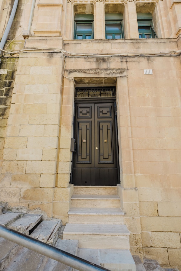 A building with a dark brown double door, stone steps leading up to it, and a window with green shutters above.