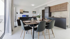 A modern kitchen with dark cabinets, a dining area with a wooden table and grey chairs, and a sofa in the background.