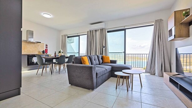 A modern living room with a grey sectional sofa, a dining area with a table and chairs, and a kitchen in the background.