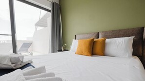 A hotel room with a large bed, white linens, two mustard pillows, and a view of a balcony with a sun lounger.