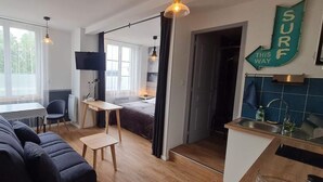 A modern living room with a sofa, a small table, and a TV. There is a kitchen area with a sink and a sign that says "SURF THIS WAY".