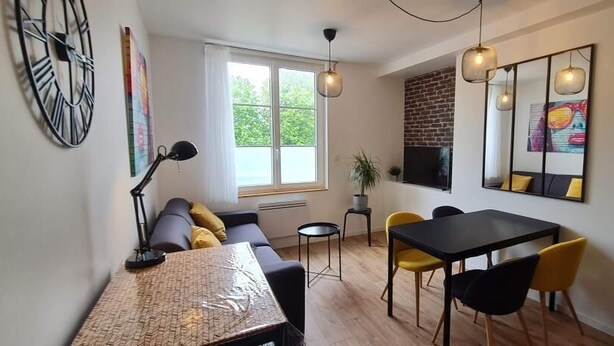 A modern living room with a sofa, dining table, and chairs. There is a window with a view of greenery outside. - Superb T1 of 28 M2 (CAEN)