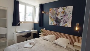 A bedroom with a bed, a desk, and a chair. There is a window with curtains, a painting on the wall, and a light switch. - Superb T1 of 28 M2 (CAEN)