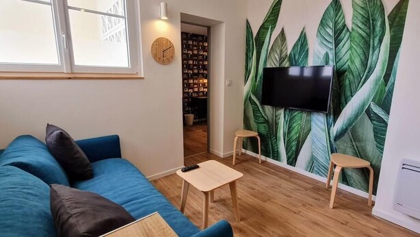 Ein gemütliches Wohnzimmer mit einem blauen Sofa, einem kleinen Holztisch, einem an einer wand mit Blätermuster befestigten Fernseher und einer Wanduhr.
