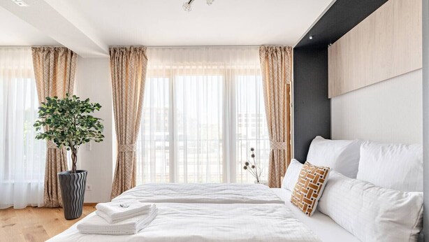 A modern bedroom with a large bed, beige curtains, a potted plant, and a view of the city. - Marllon Suites – Verso Apartment (Lörrach)