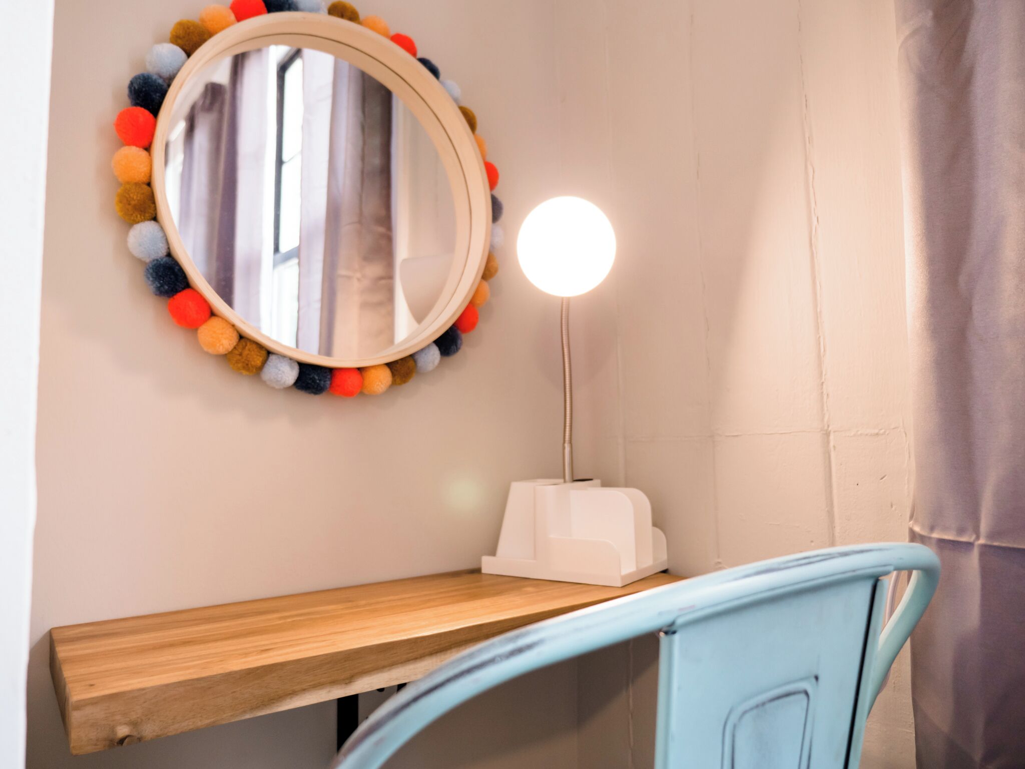 Une salle de bain avec un meuble-lavabo en bois, un miroir rond orné de pompons colorés et une lampe blanche à abat-jour rond.