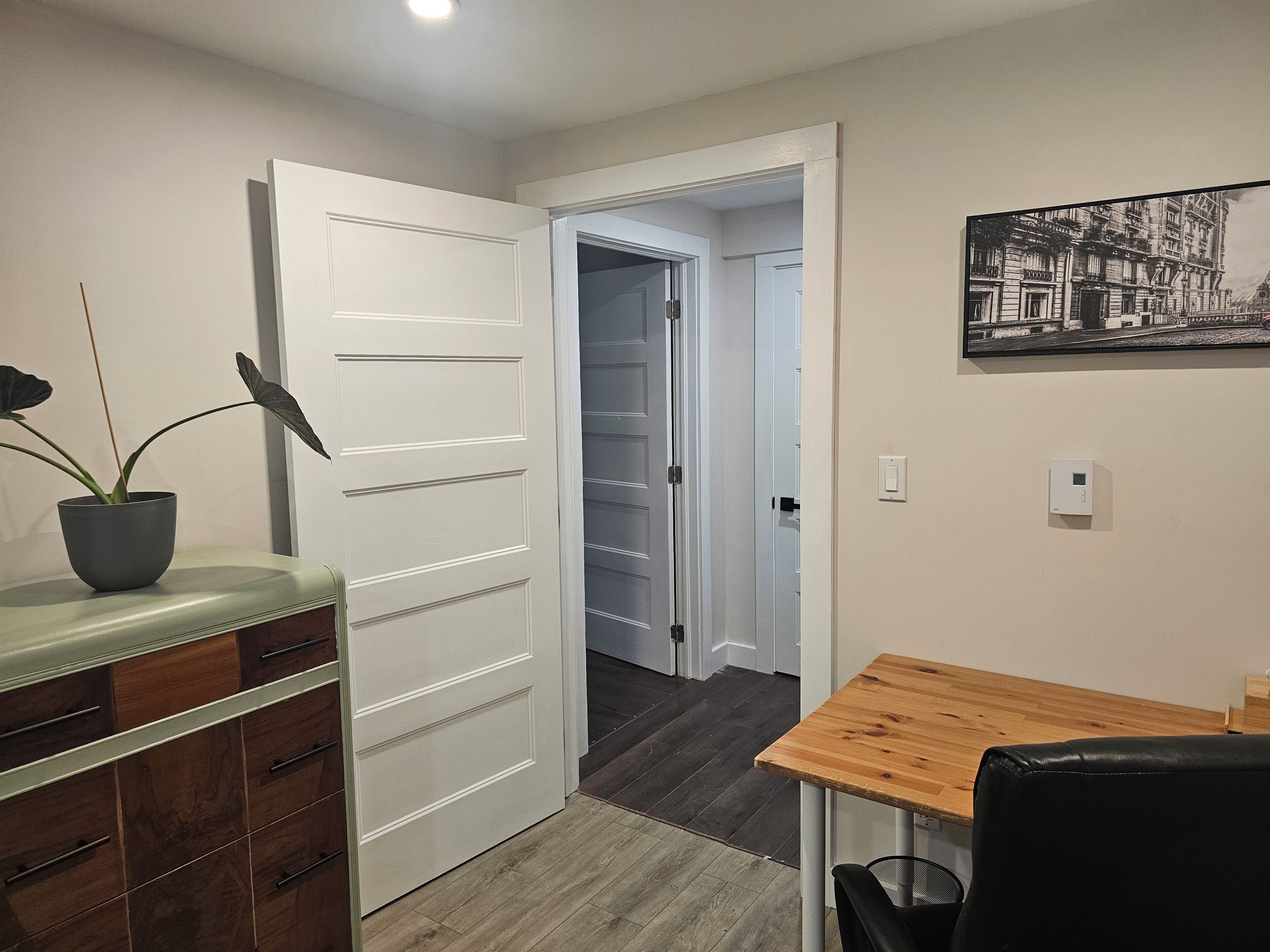 Una habitación con una mesa de comedor de madera, una planta en maceta, una puerta blanca y un pasillo que conduce a otra habitación.