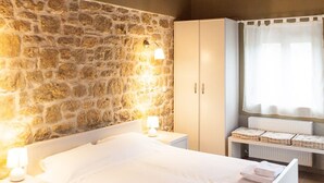 A bedroom with a stone wall, wooden ceiling beams, a bed, a wardrobe, and a window with curtains. - Villavia Residence - Sorocco Villa (West Mani)