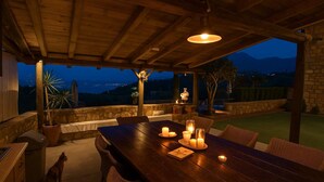 A wooden dining table with candles, wicker chairs, and a view of the mountains at dusk. - Villavia Residence - Sorocco Villa (West Mani)