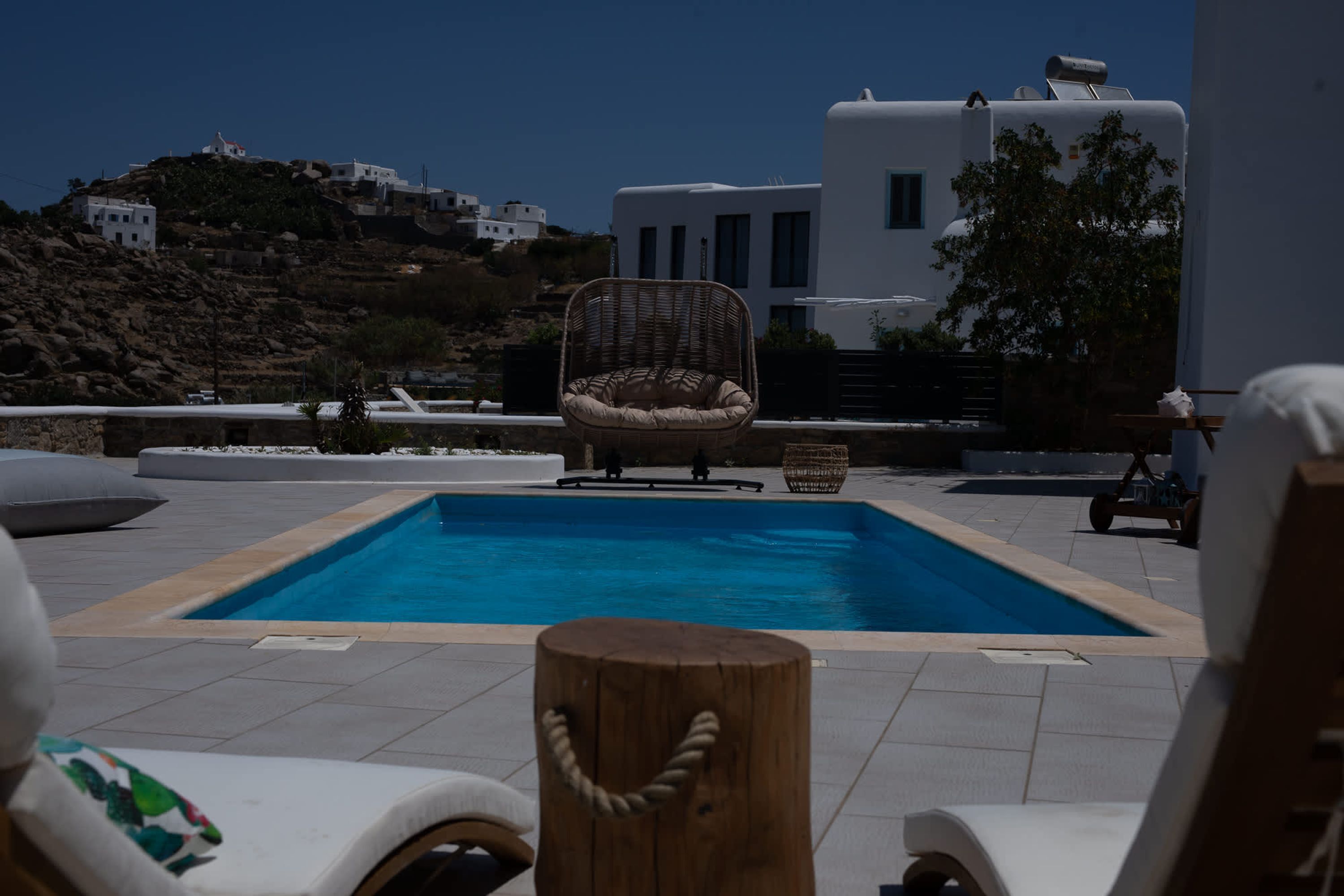 A pool area with lounge chairs and a wicker chair, surrounded by white buildings and a view of a hillside.