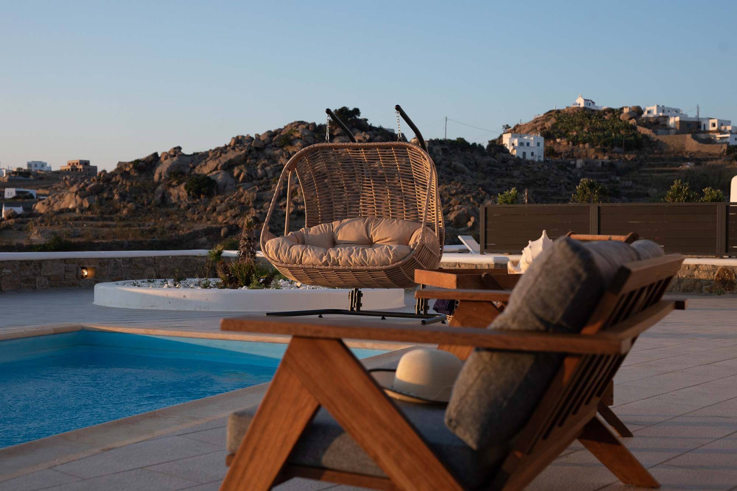 A poolside area with a wicker chair, wooden lounge chairs, and a view of a hillside with buildings.