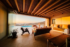 Un balcón amplio con techo de madera, zona de estar con sofá y sillones, y vistas a una piscina y el paisaje lejano.