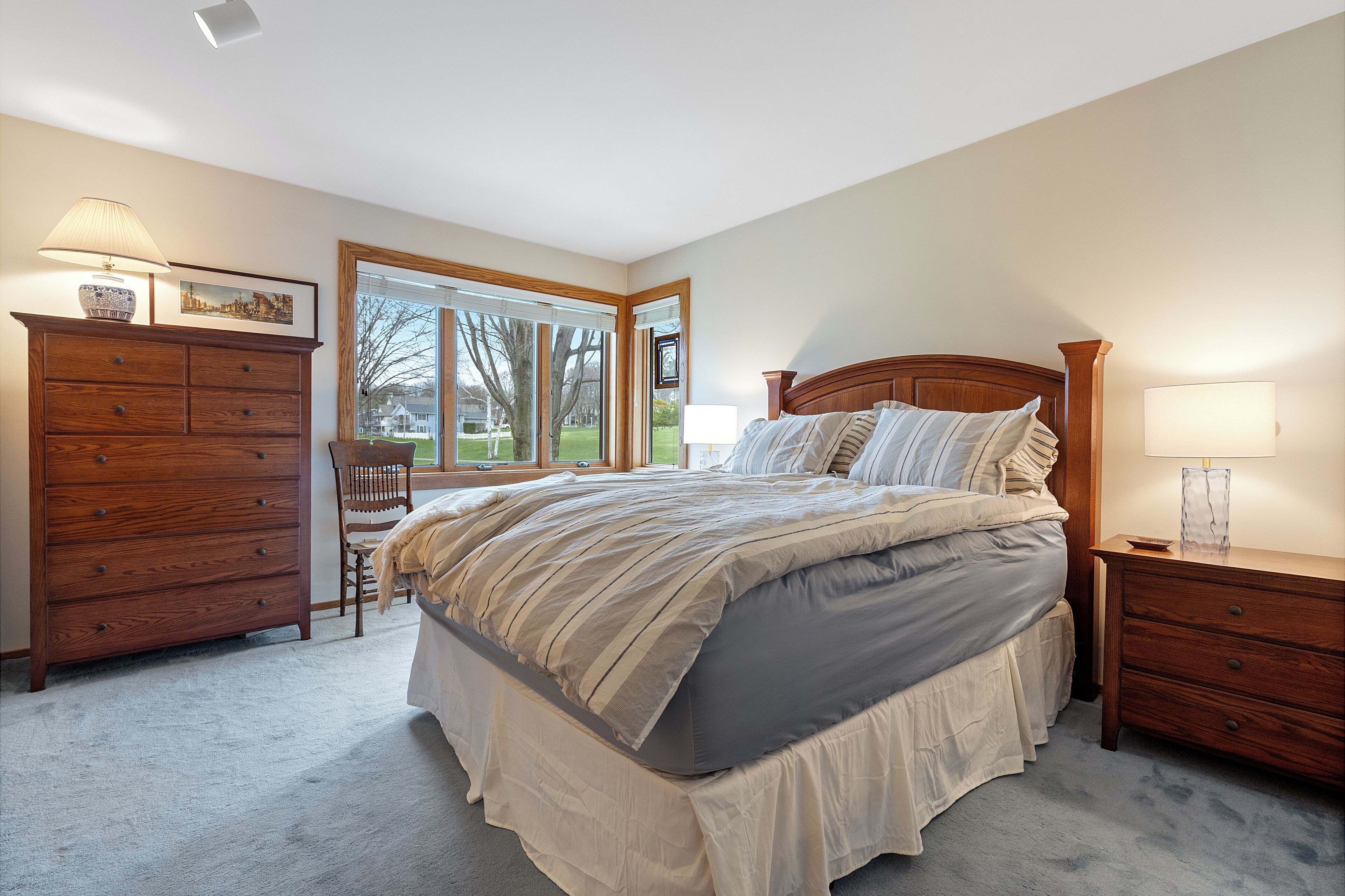 Quarto com cama grande, cômoda de madeira, cadeira e janela com vista para o exterior.